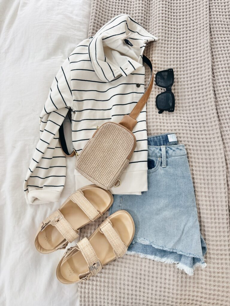 Casual spring outfit flatlay with a striped pullover, denim cutoff shorts, woven crossbody bag, and raffia sandals.