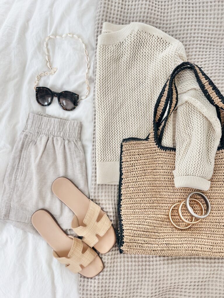 Neutral summer outfit flatlay featuring a breezy knit sweater, linen shorts, woven tote bag, raffia slide sandals, and tortoise shell sunglasses. A perfect mix of textures for a relaxed, beach-ready look.